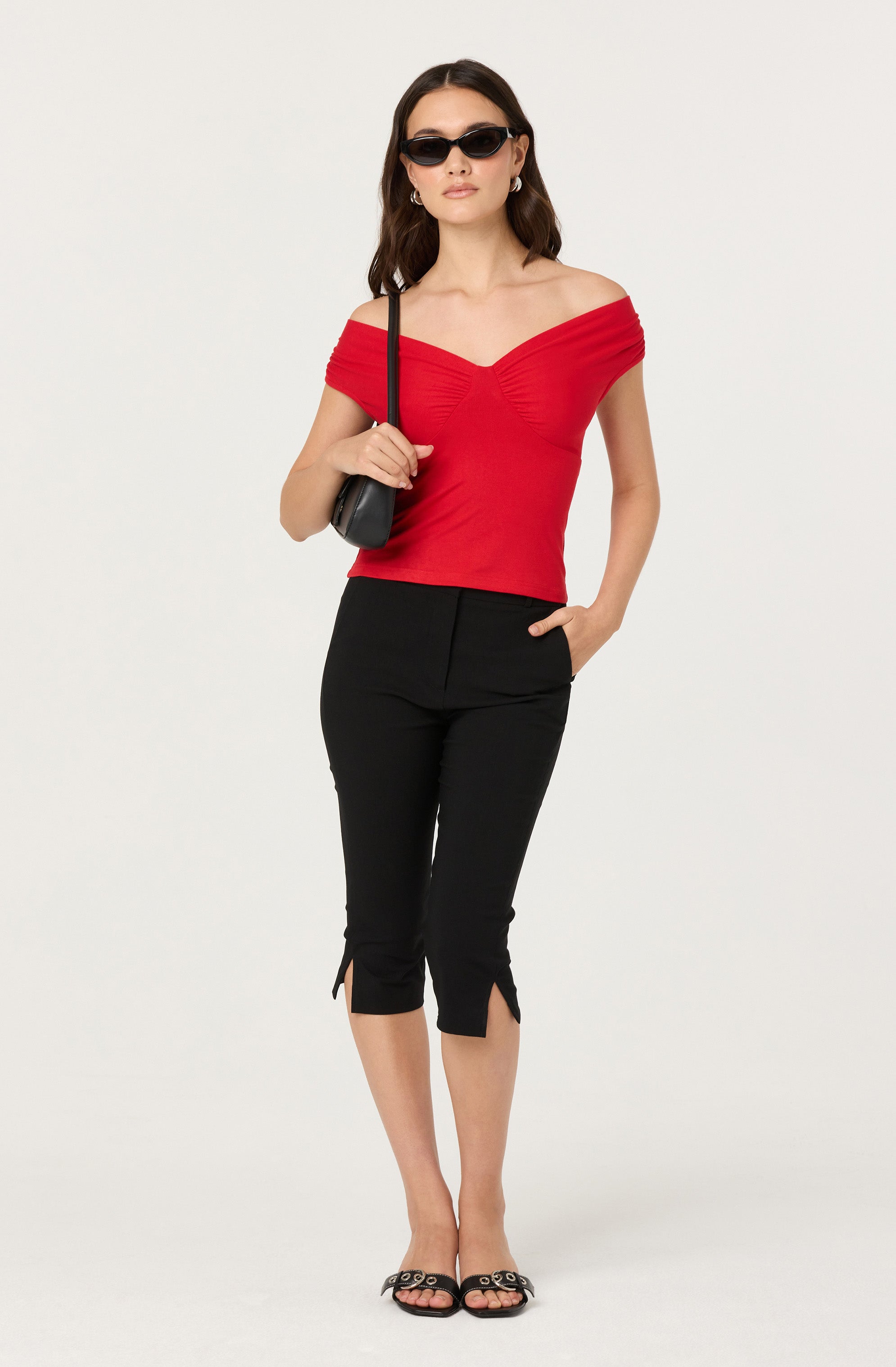 A woman is wearing a red off-the-shoulder top with a fitted, gathered bodice. She has paired it with black capri pants featuring small slits at the hems. She accessorizes with dark sunglasses, hoop earrings, and carries a small black shoulder bag. She is also wearing black open-toe sandals.