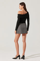 A woman is wearing an off-the-shoulder black long-sleeve top with a draped neckline, paired with a high-waisted gray plaid mini skirt. She has on black patent leather high-heeled shoes with a block heel and ankle strap. She accessorizes with gold hoop earrings and gold rings.