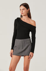A woman is wearing a black, long-sleeved, off-the-shoulder top with a gathered detail on one shoulder. She is also wearing a high-waisted, gray plaid mini skirt with front pockets.