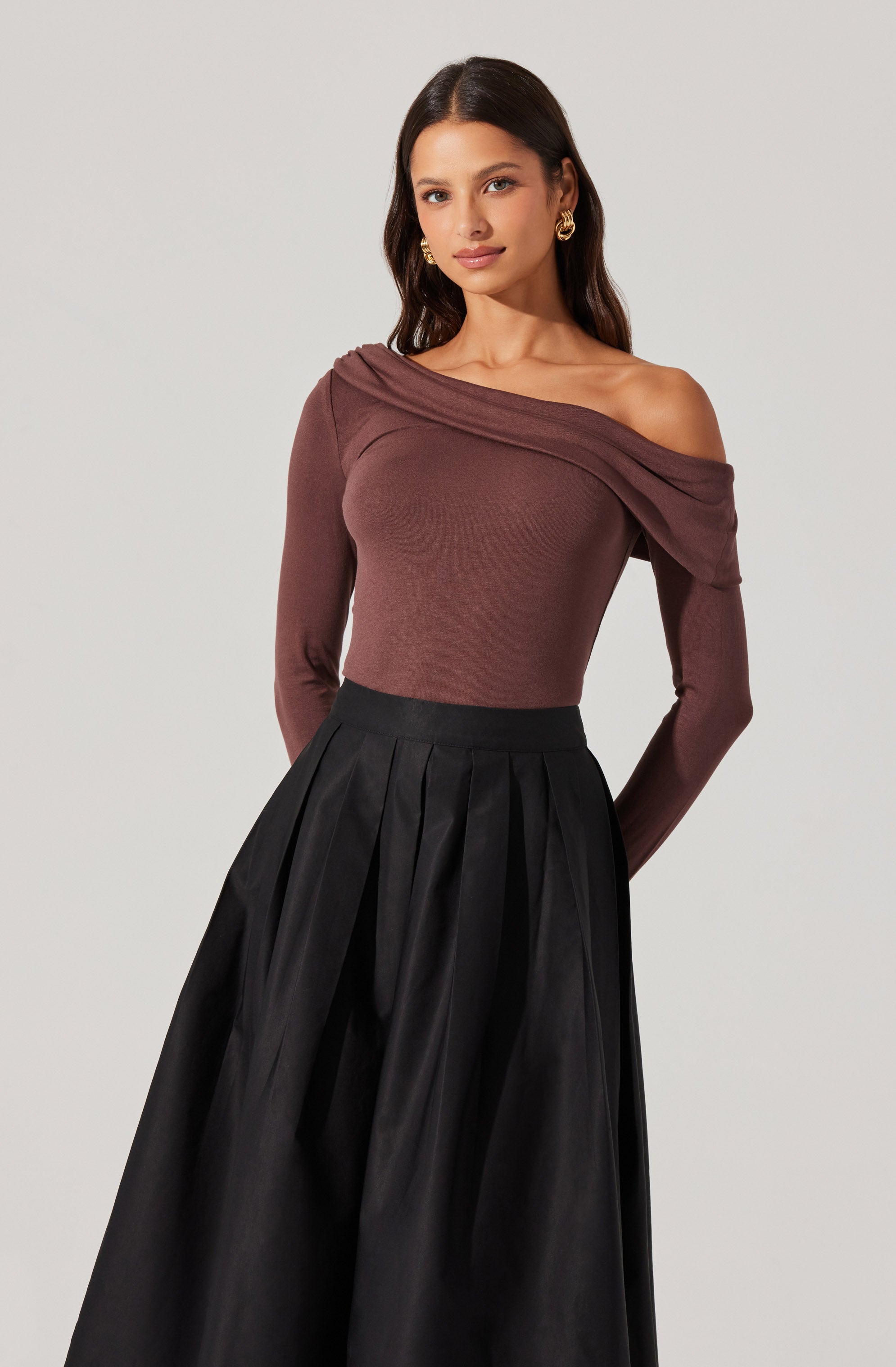 A woman is wearing a fitted, long-sleeved, off-the-shoulder top in a rich brown color, with a draped neckline that exposes her left shoulder. She pairs it with a high-waisted, full-length black skirt featuring pleats and a shiny fabric finish.