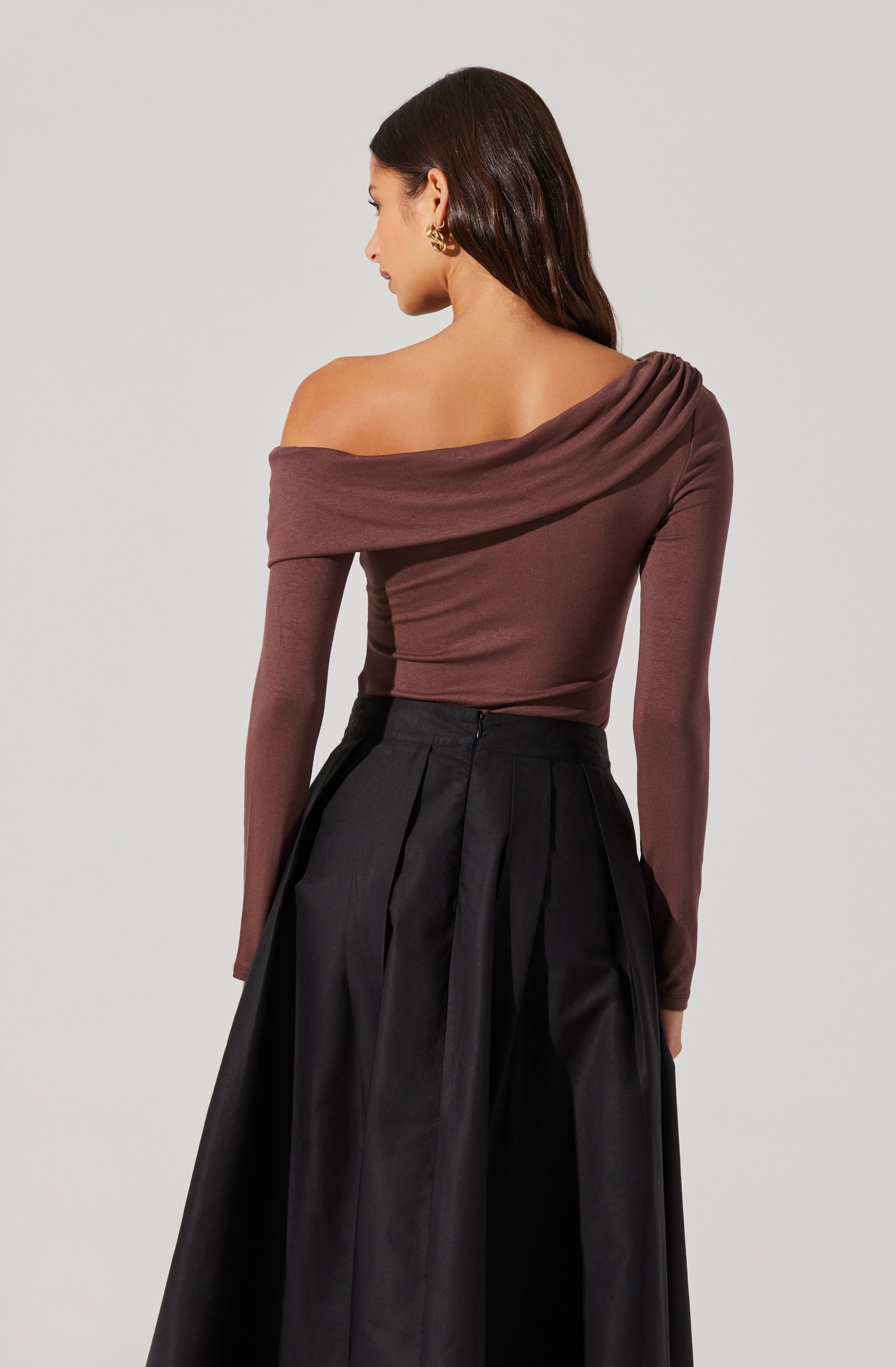 A woman is wearing an off-the-shoulder, long-sleeve top in a muted mauve or dusty pink color. The top has a draped neckline that falls over one shoulder, creating an asymmetrical look. She pairs it with high-waisted, wide-leg black trousers that have pleats running down the front. The overall outfit is stylish and sophisticated.
