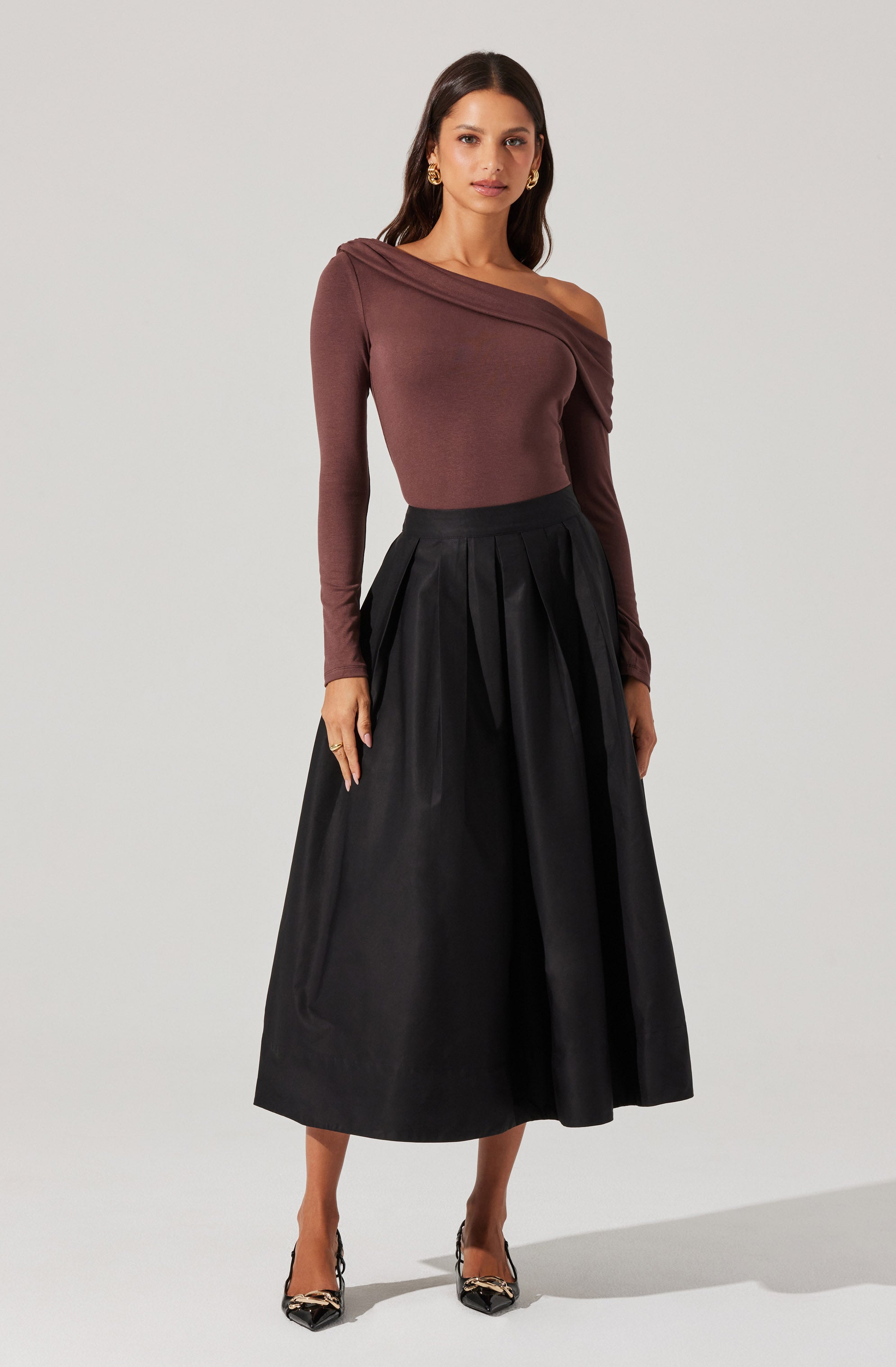 A woman is wearing a one-shoulder, long-sleeve top in a muted brown color, with the fabric draping diagonally across the shoulder. She pairs it with a high-waisted, black, full-length skirt featuring pleats and a slightly shiny finish. She completes the outfit with pointed-toe high heels that have a snakeskin pattern.