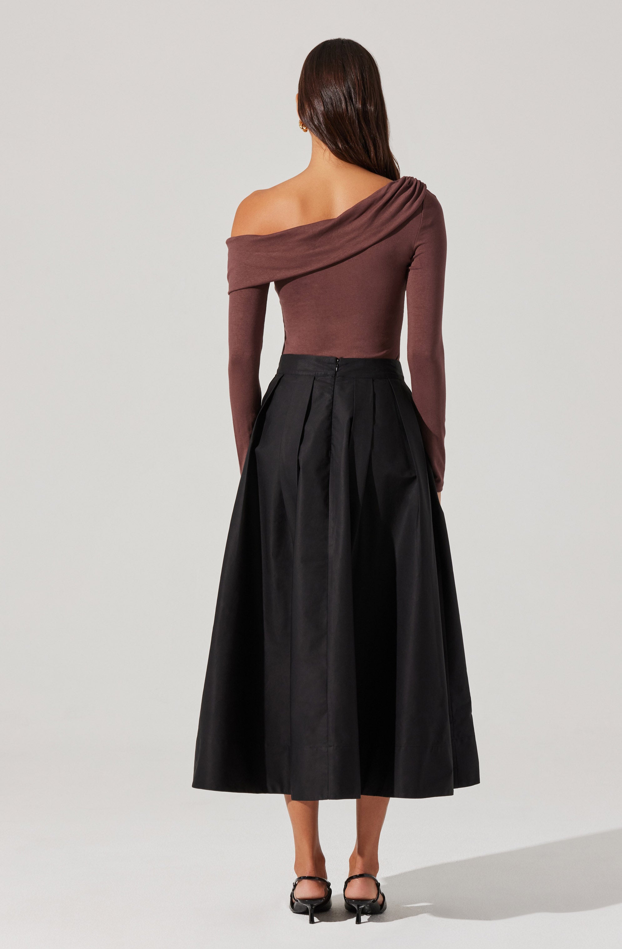 A woman is wearing a fitted, off-the-shoulder, long-sleeve top in a muted pinkish-brown color. She pairs it with a high-waisted, full-length black skirt that features pleats and a slight sheen. She completes the outfit with black open-toe heels with ankle straps.