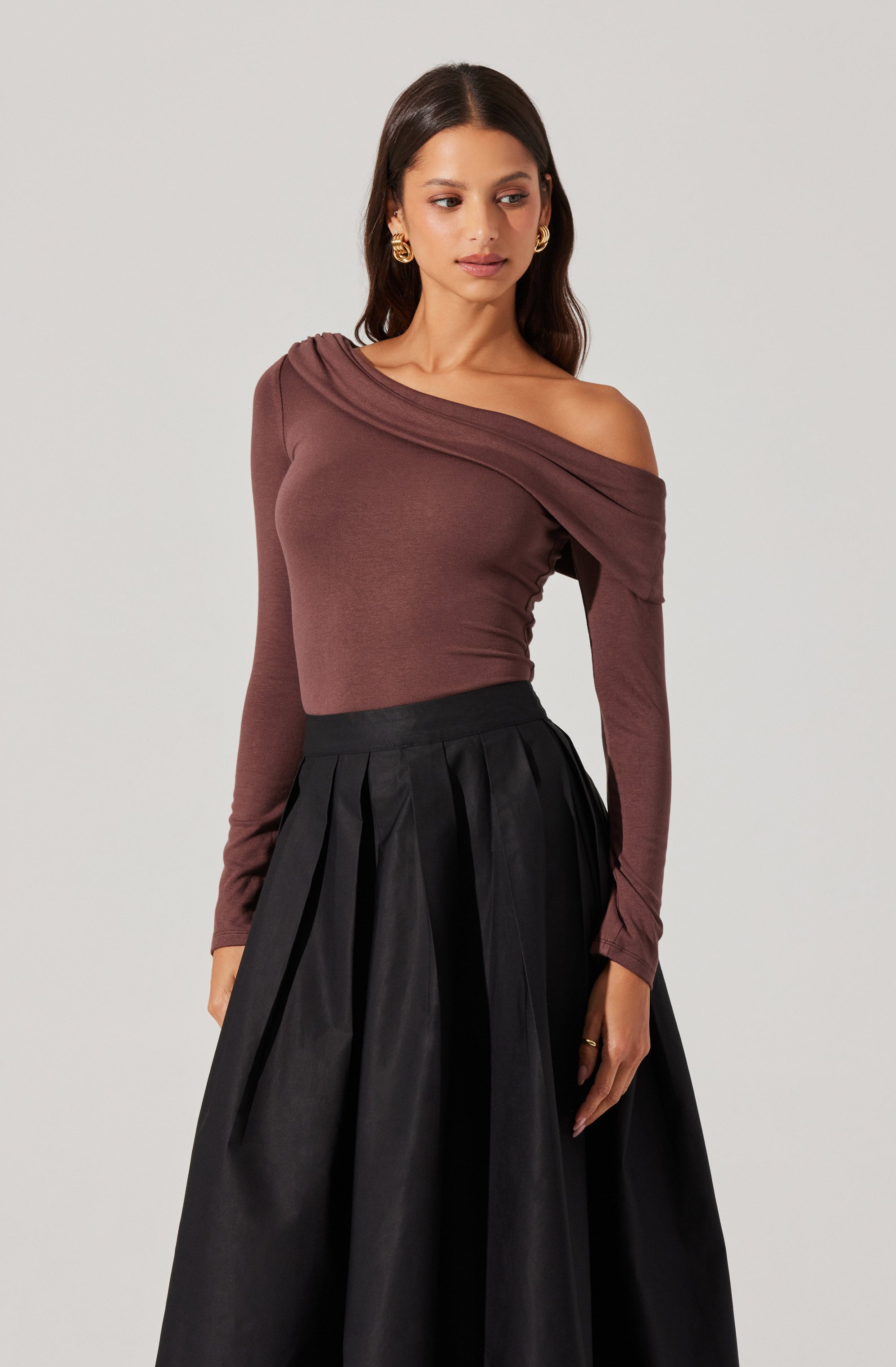 A woman is wearing a fitted, long-sleeved, off-the-shoulder top in a mauve color, with the neckline draping across one shoulder. She is also dressed in a high-waisted, full-length black skirt with pleats that create a voluminous silhouette.
