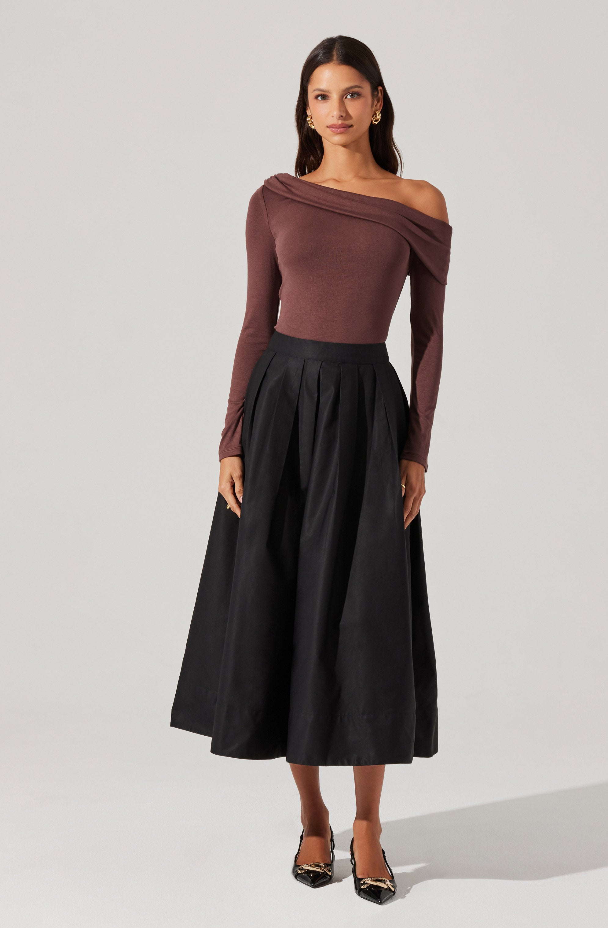 A woman is wearing an off-the-shoulder, long-sleeve top in a deep burgundy color with a draped neckline that falls to one side. She pairs it with a high-waisted, black, pleated midi skirt with a wide waistband. She completes her outfit with black pointed-toe heels featuring a decorative embellishment on the front.