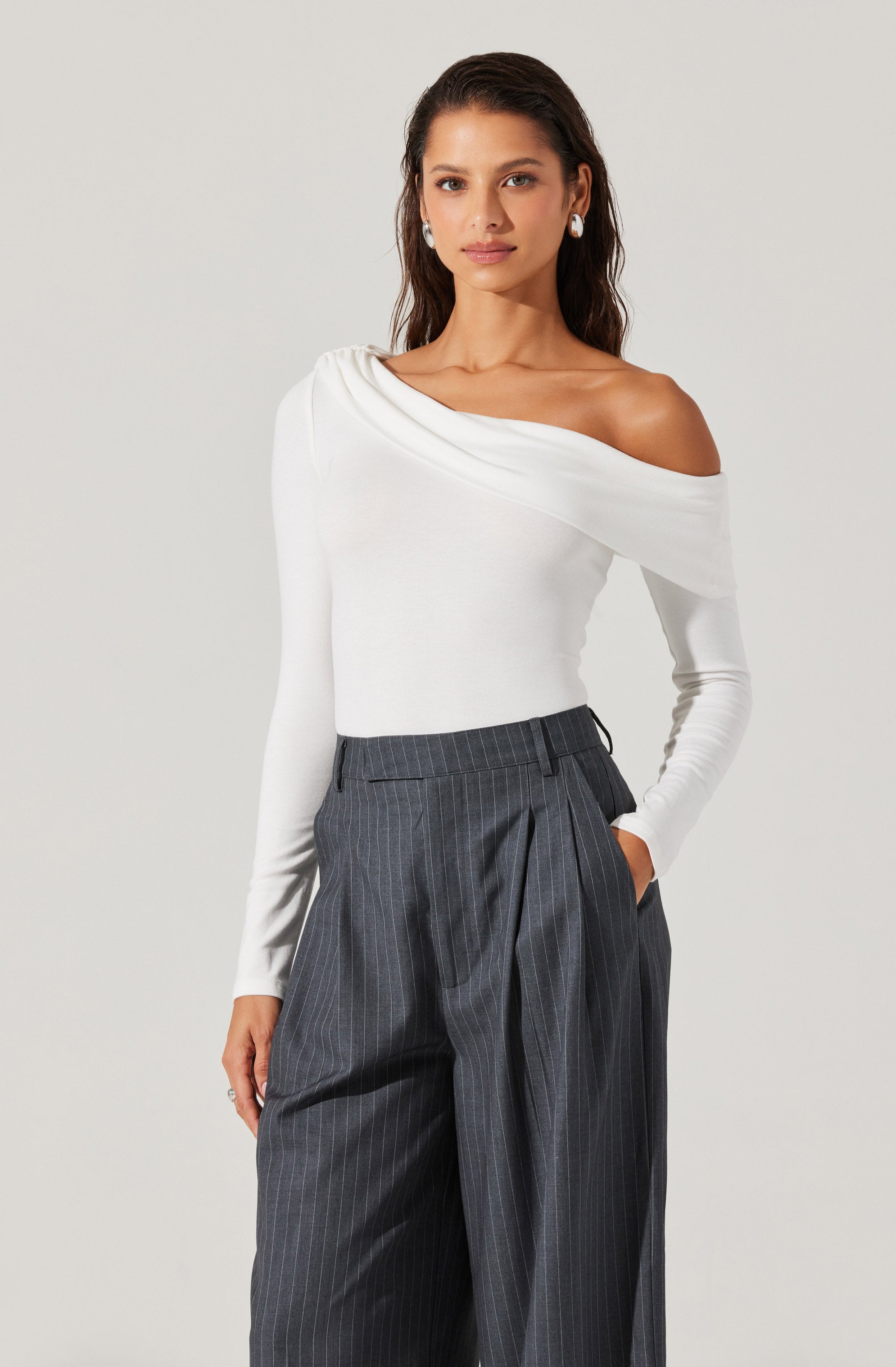 A woman is wearing a white, off-the-shoulder long-sleeve top with a draped neckline and fitted waist. She pairs it with high-waisted, dark gray pinstripe trousers featuring pleats and side pockets. The outfit is elegant and stylish, suitable for formal or professional settings.