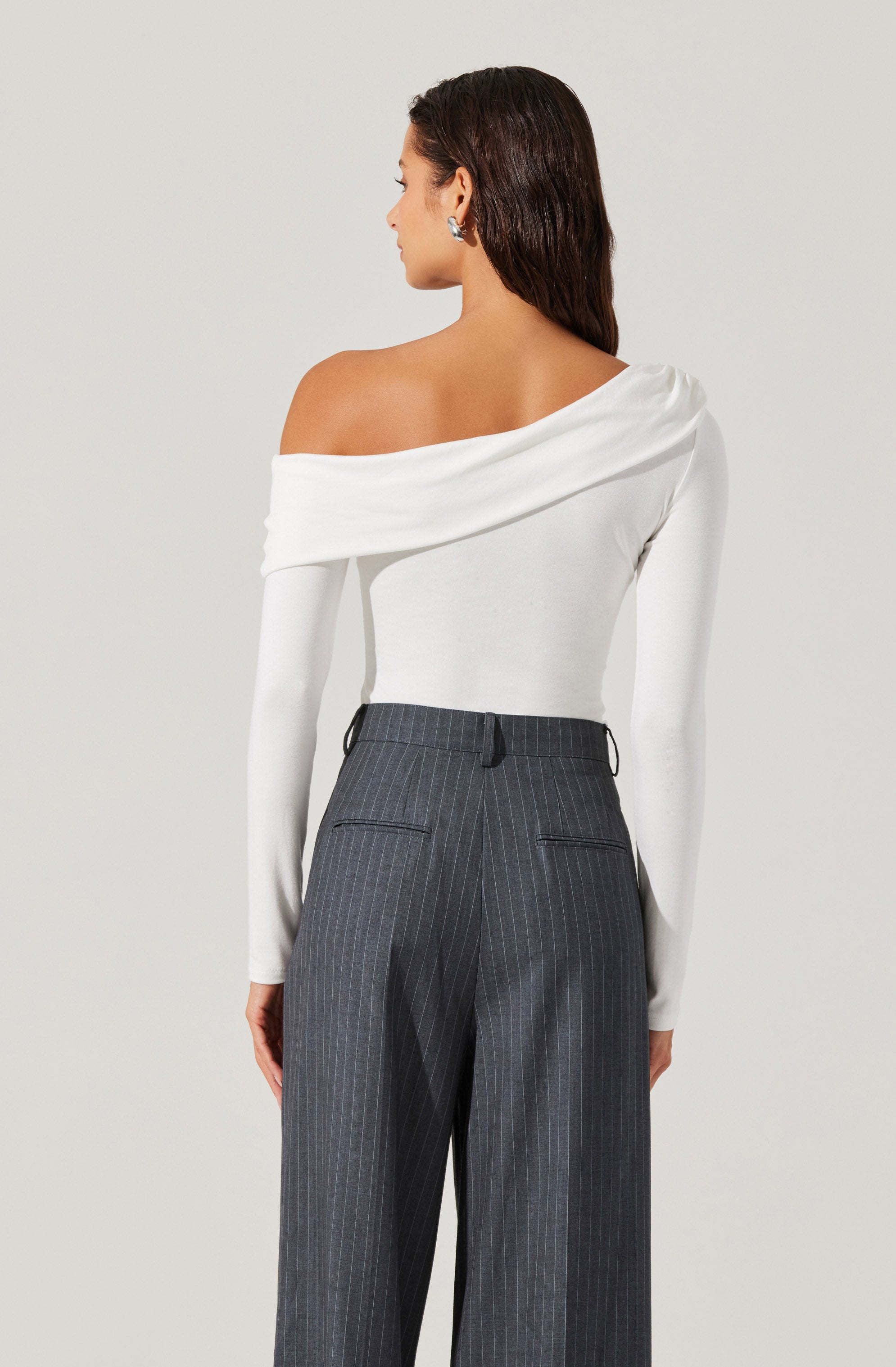 A woman is wearing an elegant, off-the-shoulder white top with long sleeves. The top has a draped detail that falls across one shoulder, creating an asymmetrical neckline. She pairs the top with high-waisted, dark gray pinstriped trousers that feature a tailored fit and belt loops.