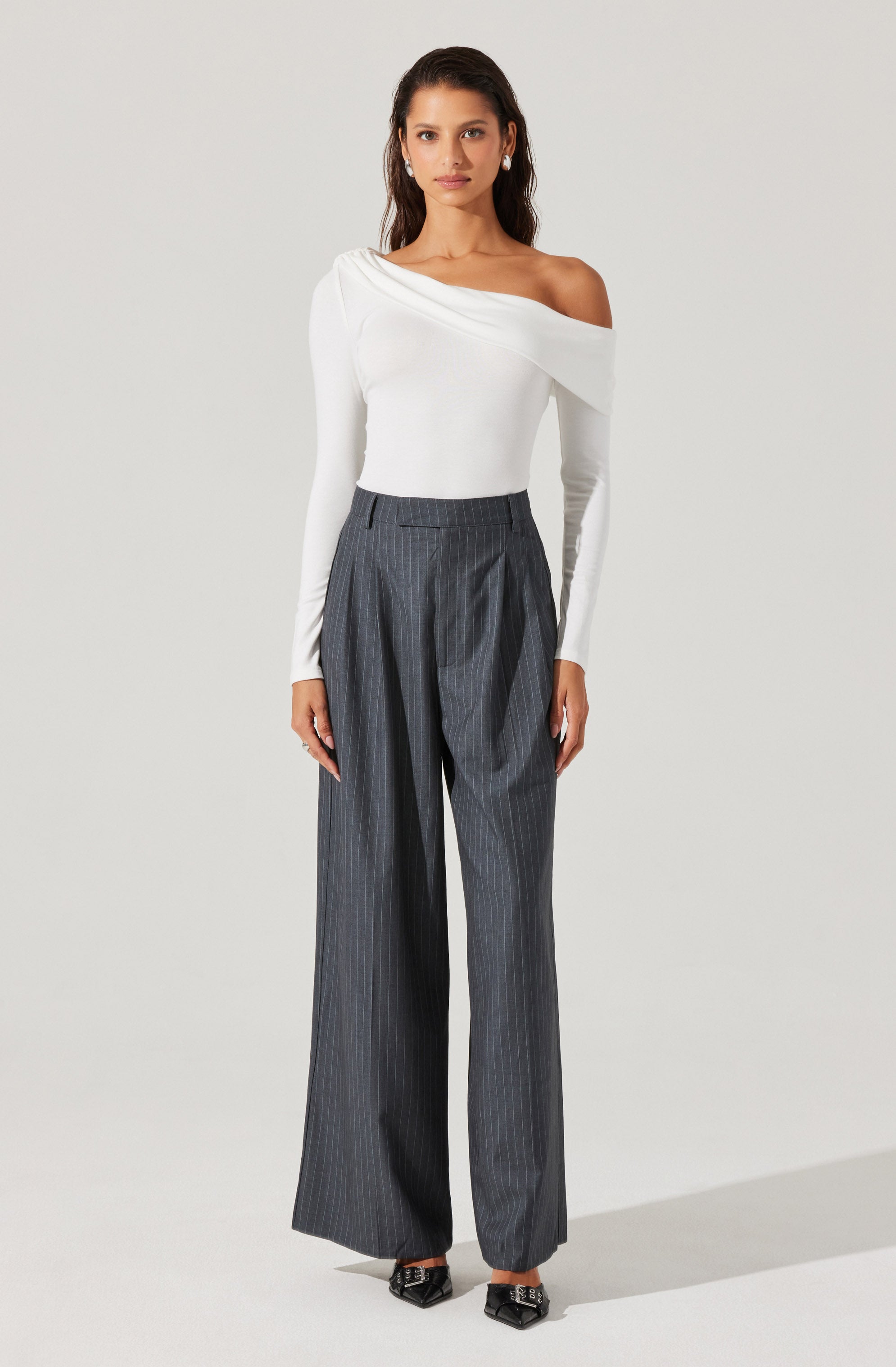 A woman is wearing a white off-the-shoulder top with long sleeves and a draped neckline, paired with high-waisted, wide-leg gray pinstripe trousers. She also has on black pointed-toe shoes with decorative buckle details.