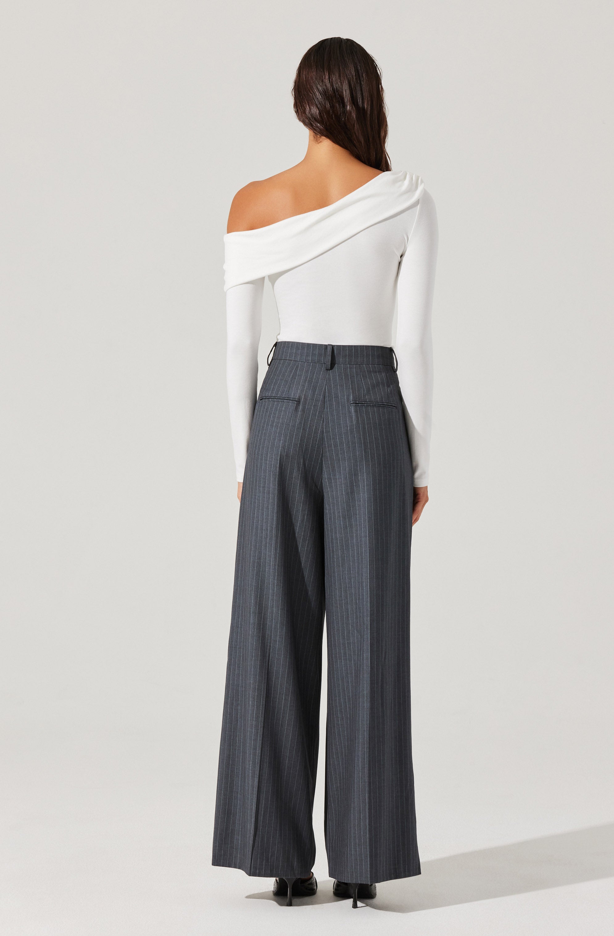 A person is wearing a white off-the-shoulder long-sleeve top with a loose drape on one shoulder, paired with high-waisted, wide-leg gray pants with pinstripes and back welt pockets.