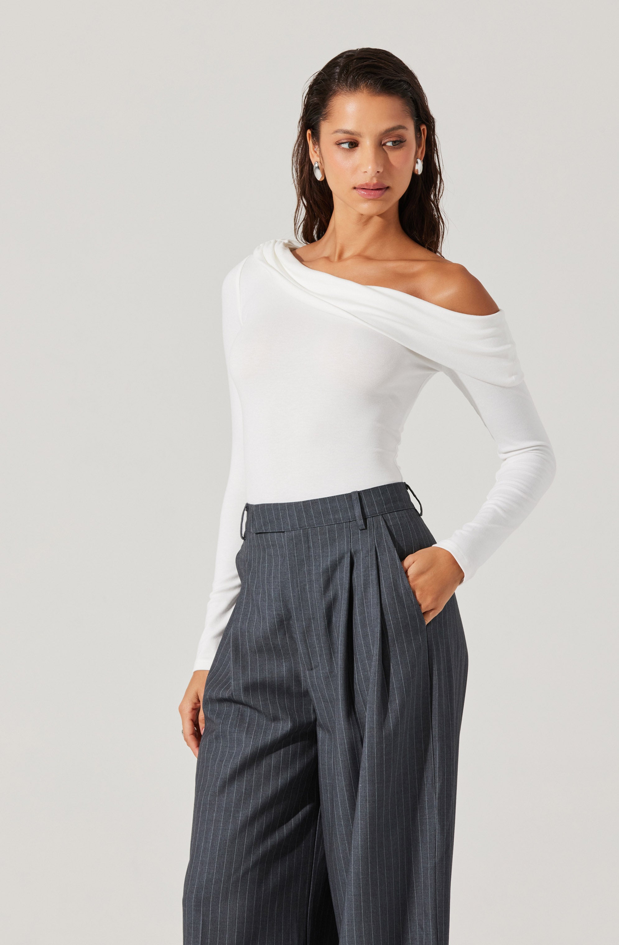 A woman is wearing a white off-the-shoulder long-sleeve top with a draped neckline, paired with high-waisted, dark gray pinstriped wide-leg trousers that feature pleats and belt loops.