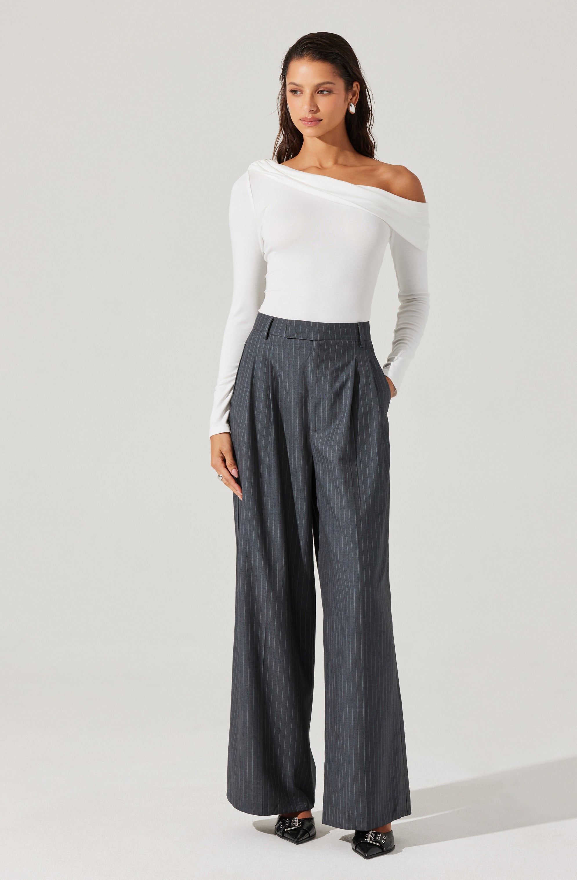 A woman is wearing a white, off-the-shoulder, long-sleeve top with a fitted silhouette. She pairs it with high-waisted, wide-leg gray trousers featuring a subtle pinstripe pattern and pleats at the front.