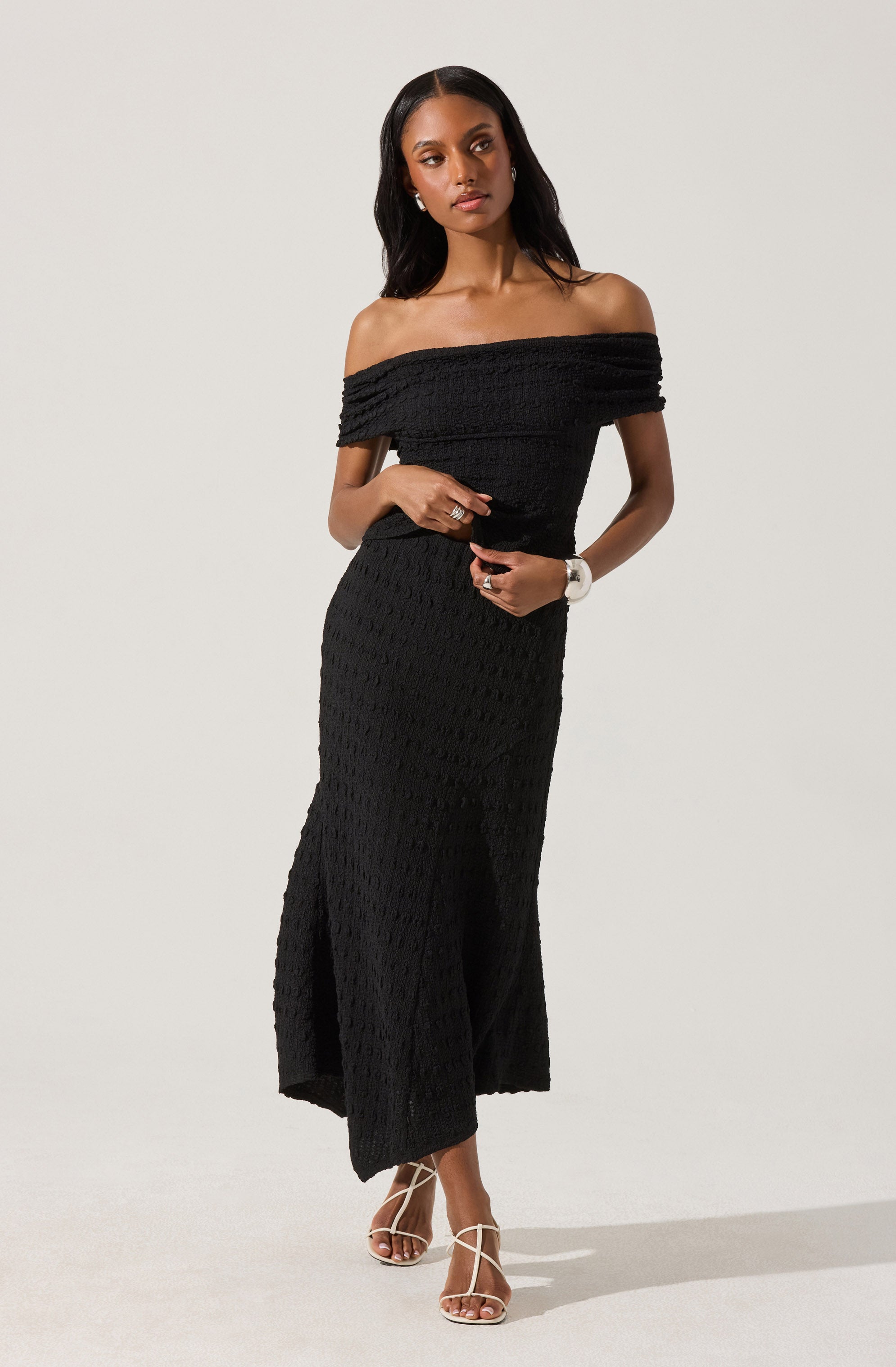 A woman is wearing a black, off-the-shoulder, knit dress that is fitted at the waist and flows into a long, slightly flared skirt with a slit on one side. She has paired the dress with strappy high-heeled sandals.