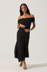 A woman is wearing a black, off-the-shoulder, knit dress that is fitted at the waist and flows into a long, slightly flared skirt with a slit on one side. She has paired the dress with strappy high-heeled sandals.