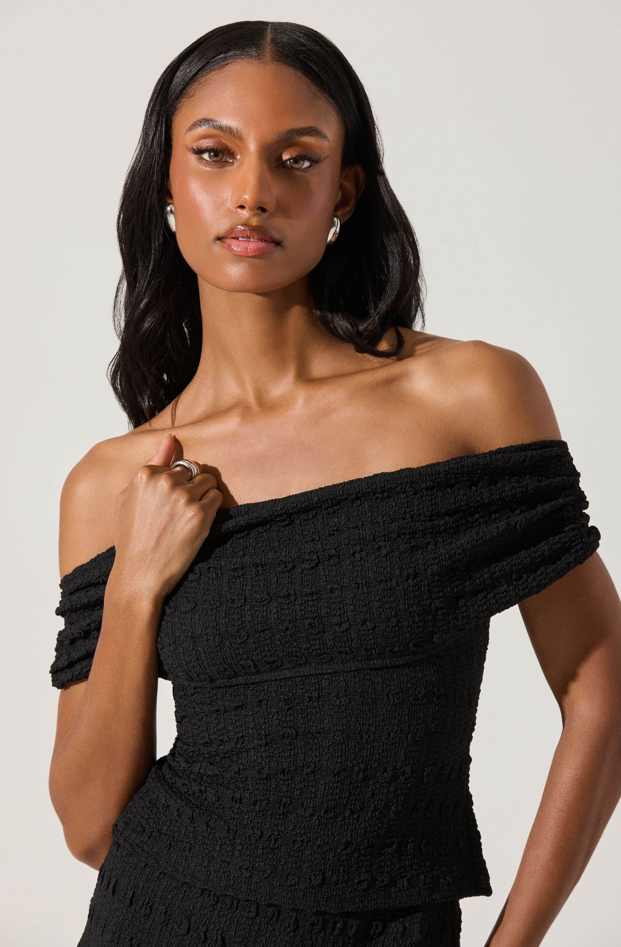 A woman is wearing an off-the-shoulder black textured dress with a fitted silhouette. The dress features short sleeves that are slightly gathered and a straight neckline that sits off the shoulders.