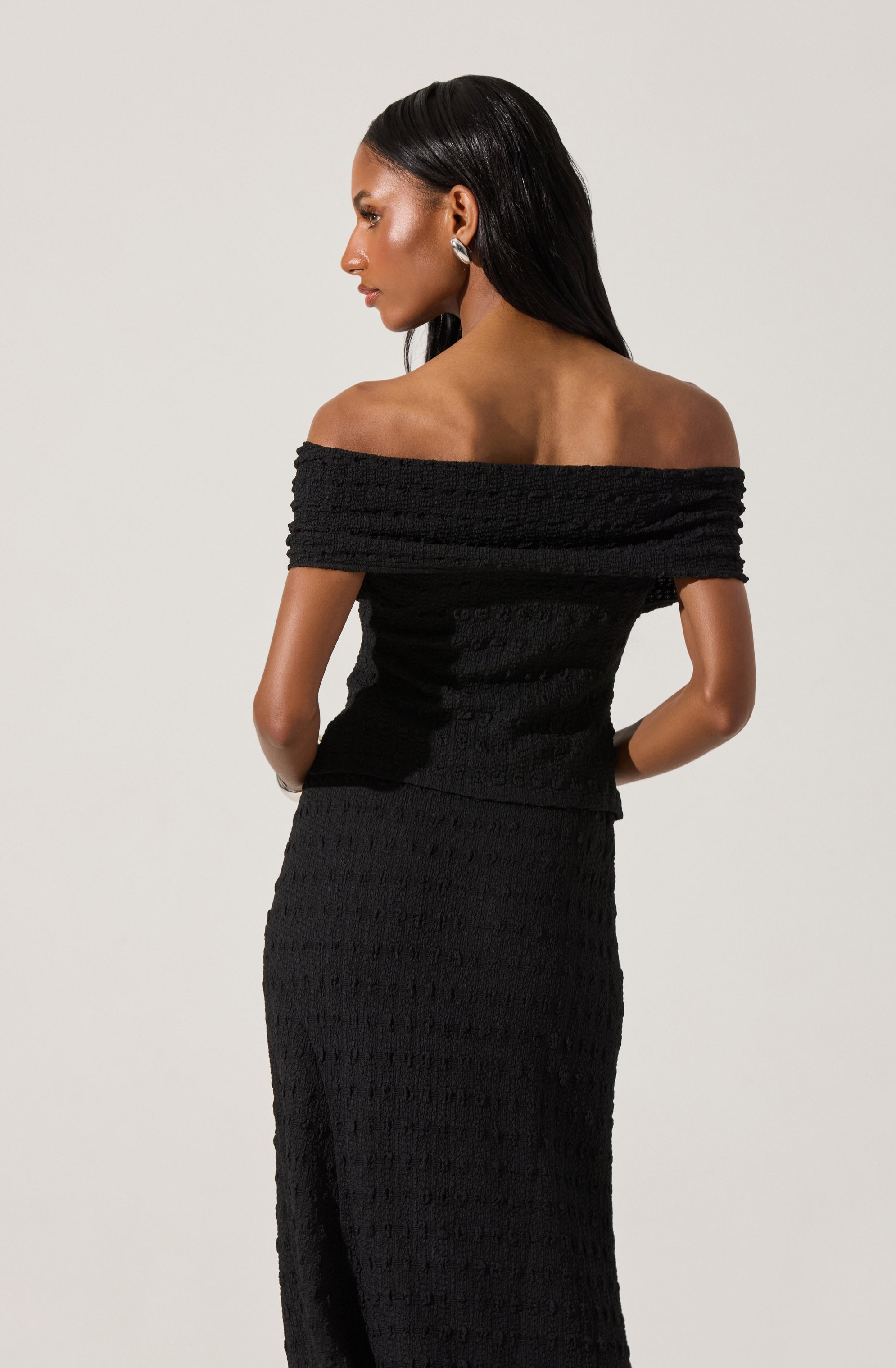 A woman is wearing a black off-the-shoulder dress with a textured, woven pattern. The dress has a wide neckline that exposes her shoulders and collarbone, and it is form-fitting, extending down to her ankles. She is accessorized with hoop earrings and has long, straight, dark hair.