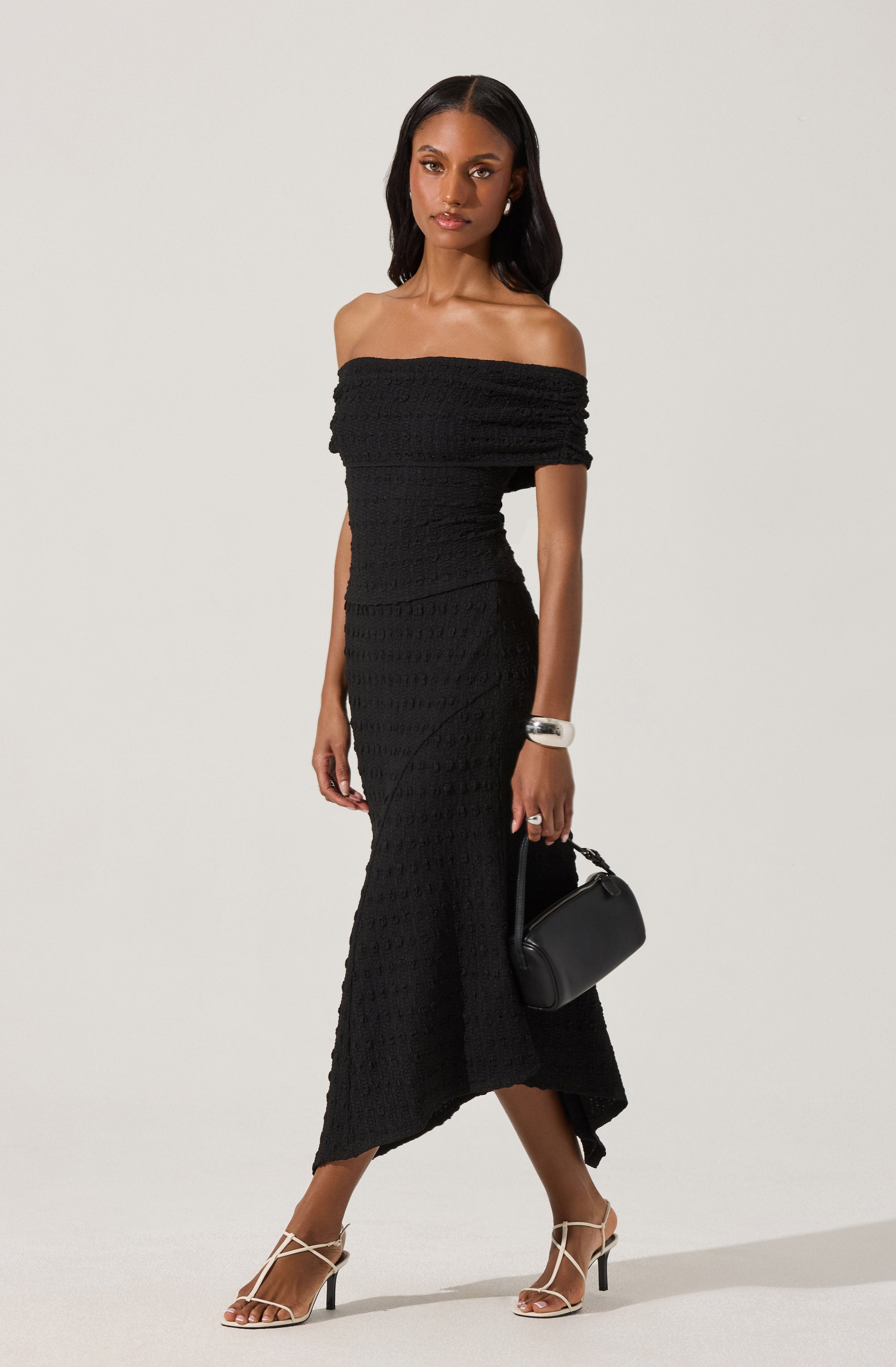 A woman is wearing an off-the-shoulder black dress with a textured, knitted appearance. The dress is fitted at the waist and extends to a midi length with a slight asymmetrical hem. She accessorizes with a silver bracelet on her right wrist, a ring on her left hand, and is holding a small black handbag. She is also wearing white high-heeled sandals with thin straps.