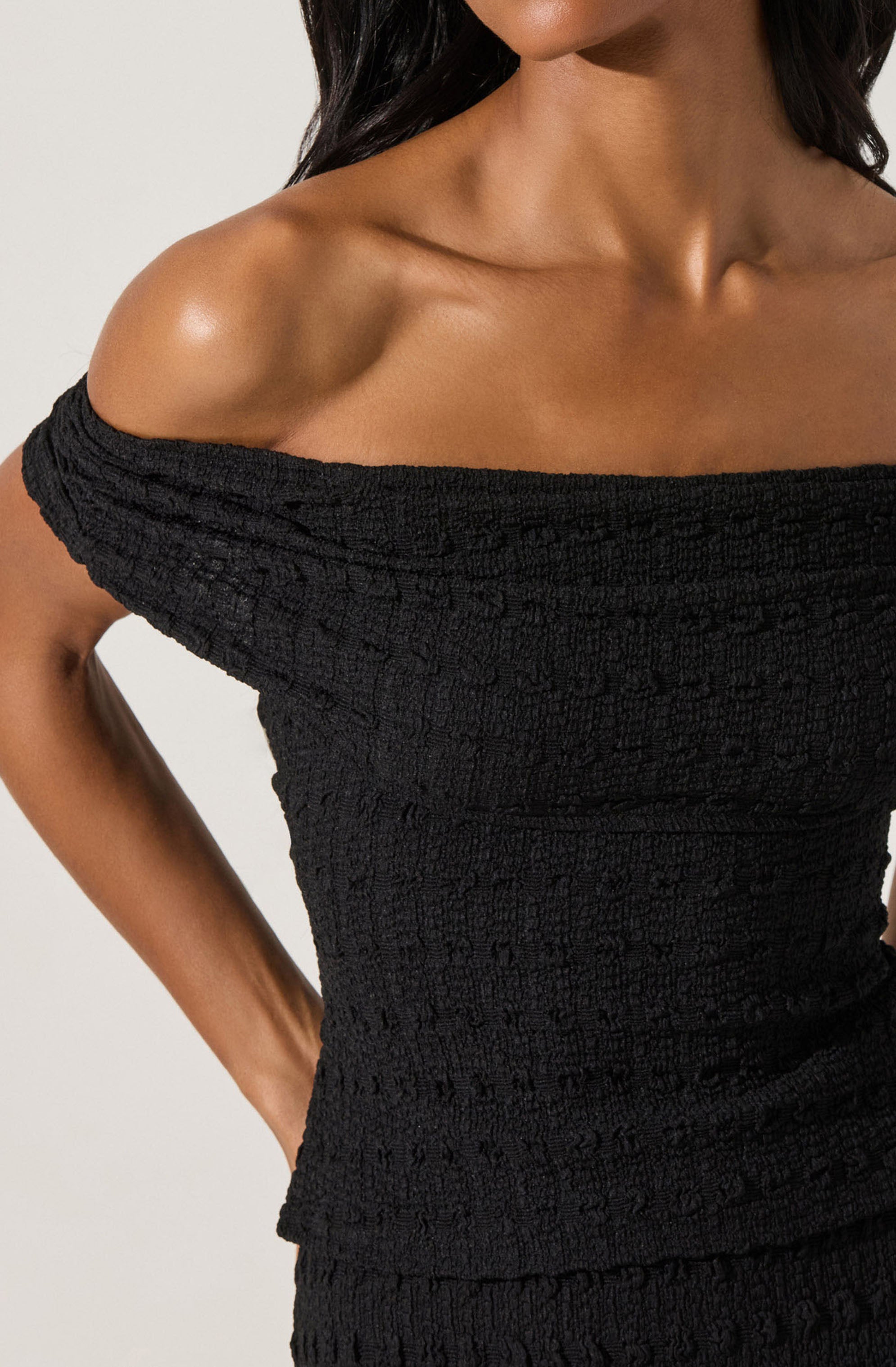 A woman is wearing an off-the-shoulder black dress with a textured, ruched fabric that fits snugly around her torso. The dress exposes her shoulders and collarbone, highlighting her skin tone.