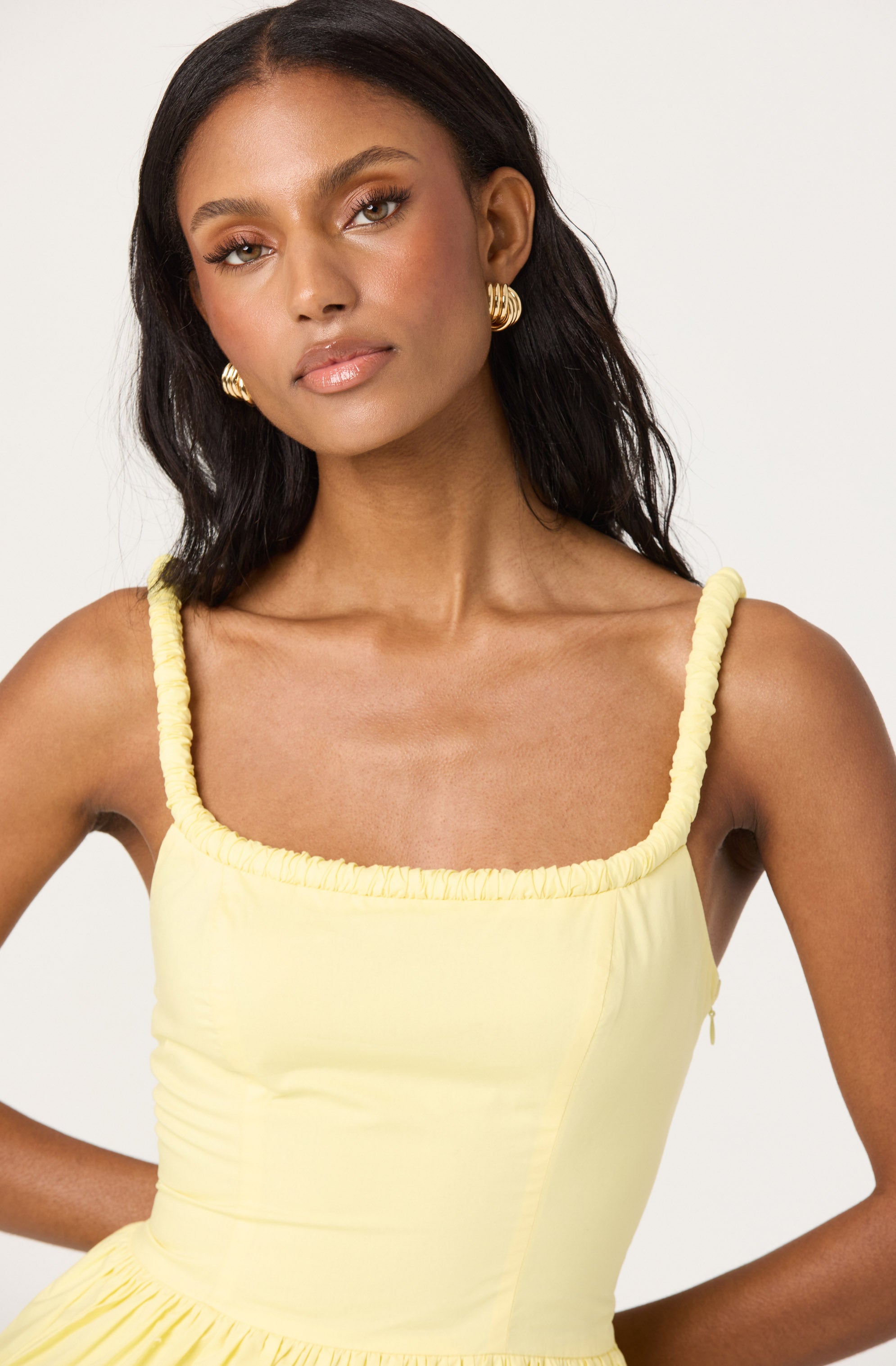 A woman with medium skin tone and long, dark hair wearing a sleeveless, pale yellow dress with gathered, ruched straps and a square neckline. She has on gold, twisted hoop earrings and a natural makeup look.