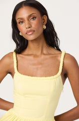 A woman with medium skin tone and long, dark hair wearing a sleeveless, pale yellow dress with gathered, ruched straps and a square neckline. She has on gold, twisted hoop earrings and a natural makeup look.