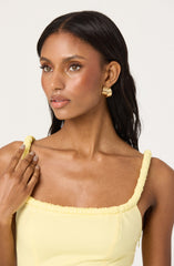 A woman is wearing a sleeveless, light yellow top with gathered fabric around the shoulder straps and neckline. She has large, gold hoop earrings and her dark hair is styled straight, parted in the middle.