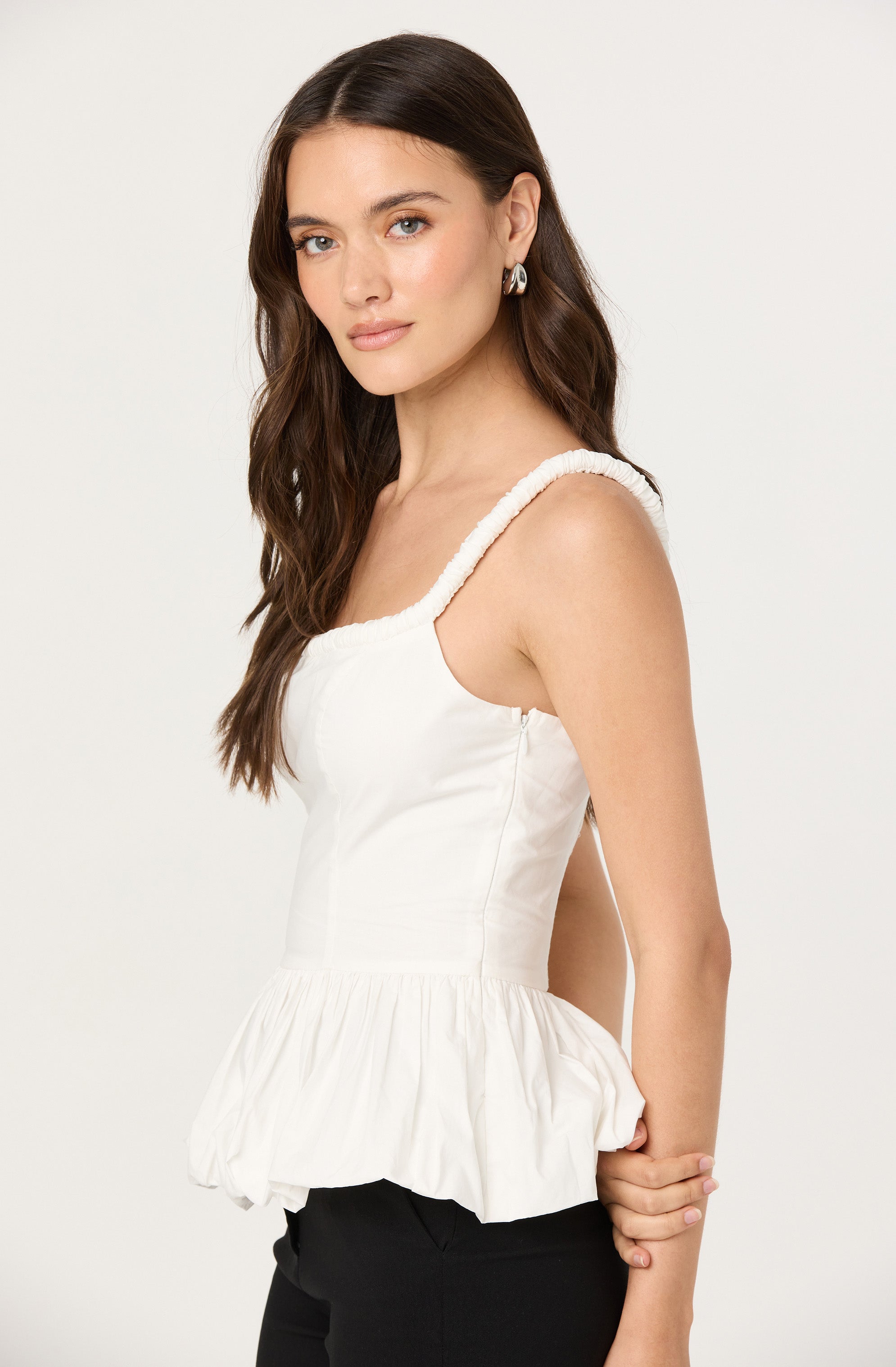 A woman is wearing a sleeveless, white top with textured straps and a gathered, ruffled hem. She pairs the top with black pants.