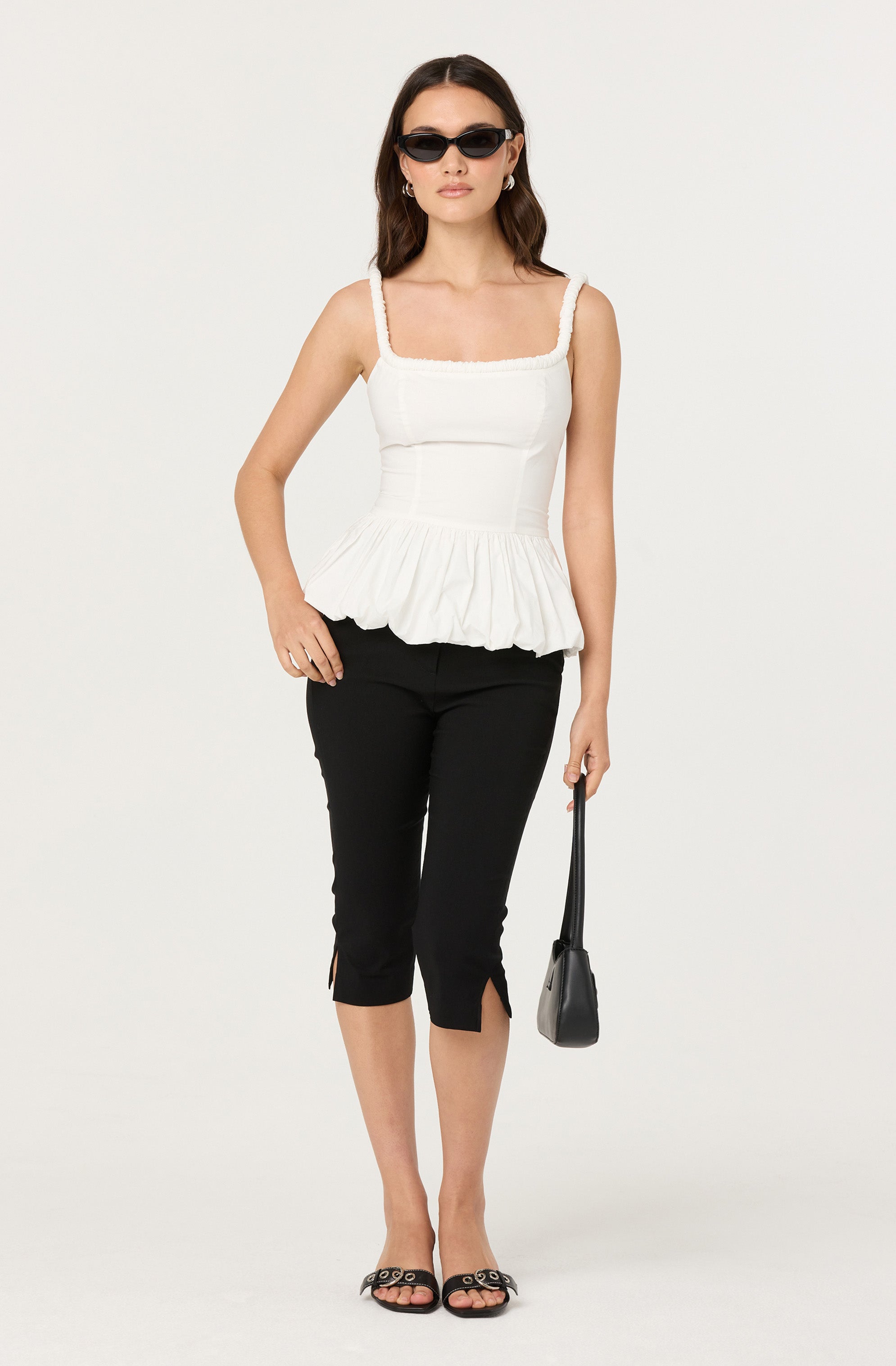 A woman is wearing a white sleeveless top with thick straps and a ruffled hem. She pairs the top with black cropped pants that have small slits at the front of each leg. She accessorizes with black sunglasses, earrings, and black open-toe sandals with grommet details. She is holding a small black handbag.