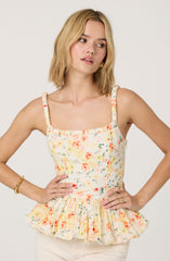 A woman is wearing a sleeveless, floral-patterned top with ruffled edges along the waist. The fabric features a light background with a colorful print of orange, pink, and yellow flowers and green leaves. The top has thin, textured straps and a square neckline. She is accessorizing with gold hoop earrings, a gold bangle on her right wrist, and multiple rings on her fingers. The overall style is feminine and summery.