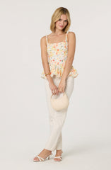 A woman is wearing a sleeveless, floral-patterned top with ruffled hem and tie straps, paired with high-waisted, cream-colored trousers. She accessorizes with a small, cream-colored handbag, gold bracelets, and rings, and is wearing white, open-toed heeled sandals.