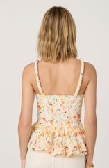 A woman is wearing a sleeveless, floral-printed top with ruffled details at the waist. The top has thin, textured straps and a fitted bodice with a smocked back. The fabric features a delicate pattern of small, colorful flowers on a light background.