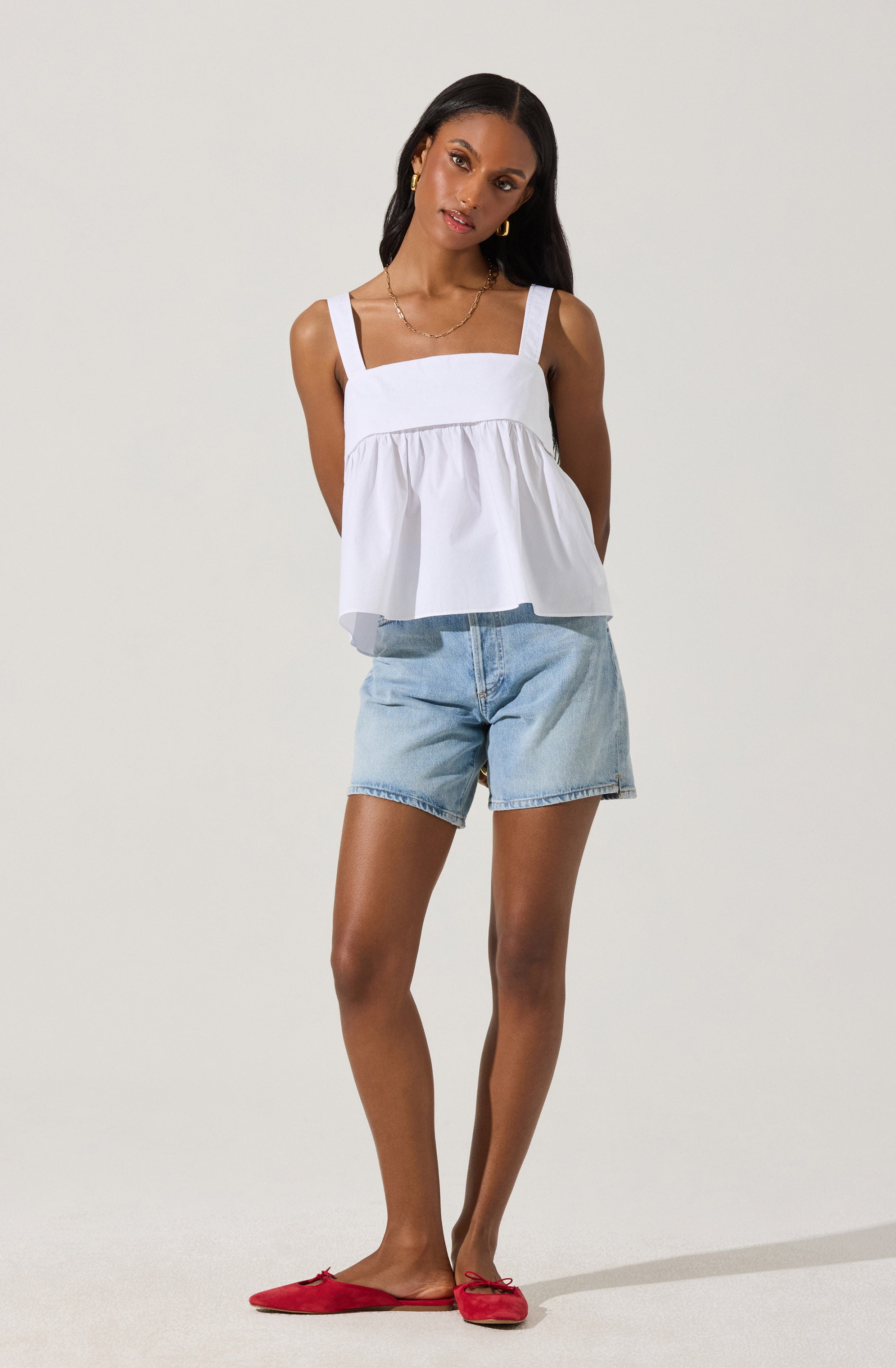 A woman is wearing a white sleeveless top with wide shoulder straps and a gathered, ruffled hem. She has on light blue denim shorts and red flat shoes with small bows on the toes.