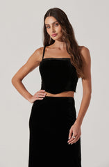 A woman is wearing a black velvet sleeveless crop top with thin straps and a high-waisted black velvet skirt.