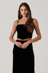 A woman is wearing a black velvet crop top with thin spaghetti straps and a high-waisted black skirt.