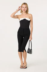 A woman is wearing a strapless black jumpsuit with a white ruffle detail across the bust. She is also wearing black high-heeled sandals and holding a small black handbag.