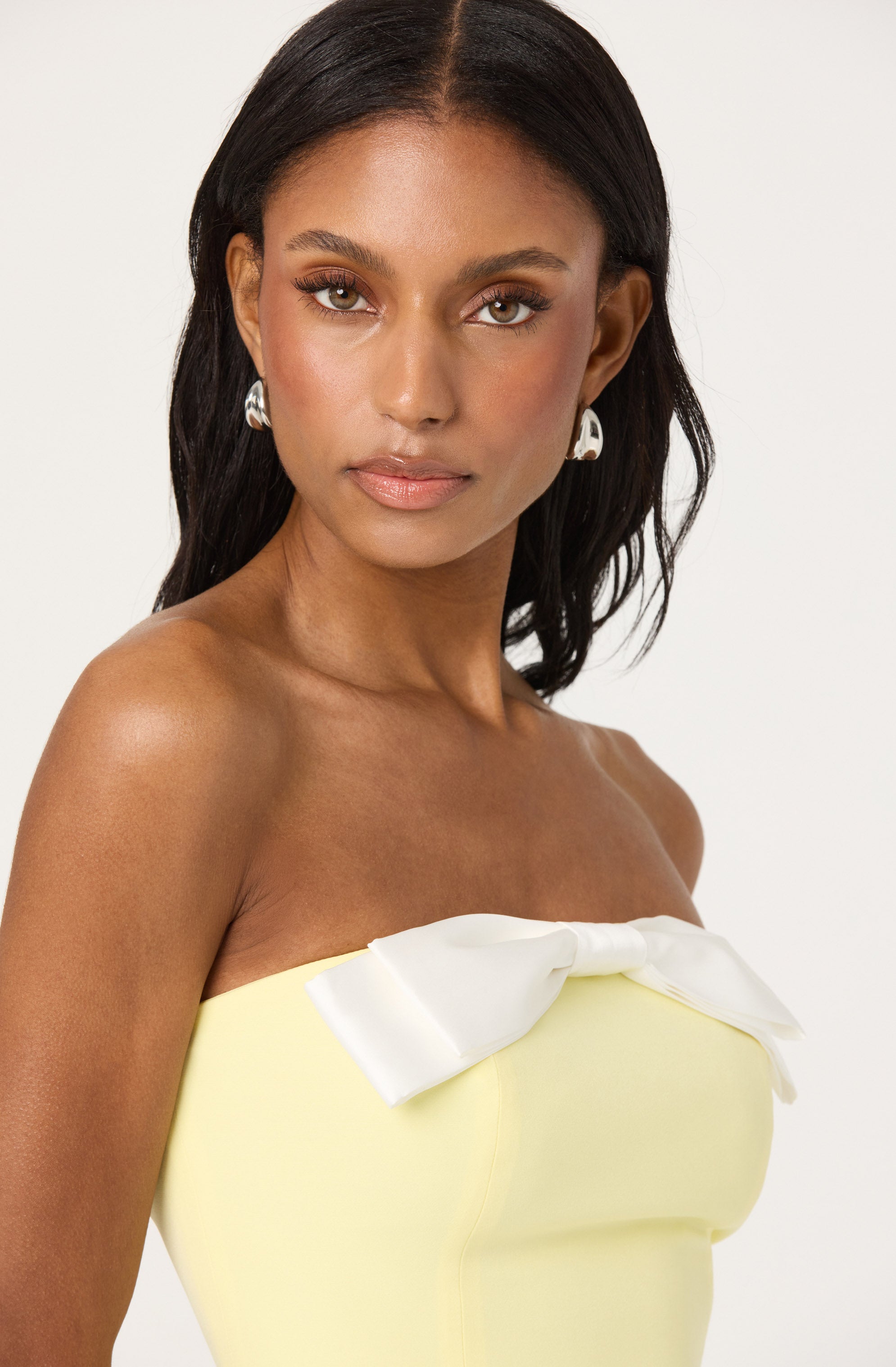 A woman is wearing a strapless, light yellow dress with a large white bow at the center of the neckline. She has on silver earrings and her hair is styled straight and loose.