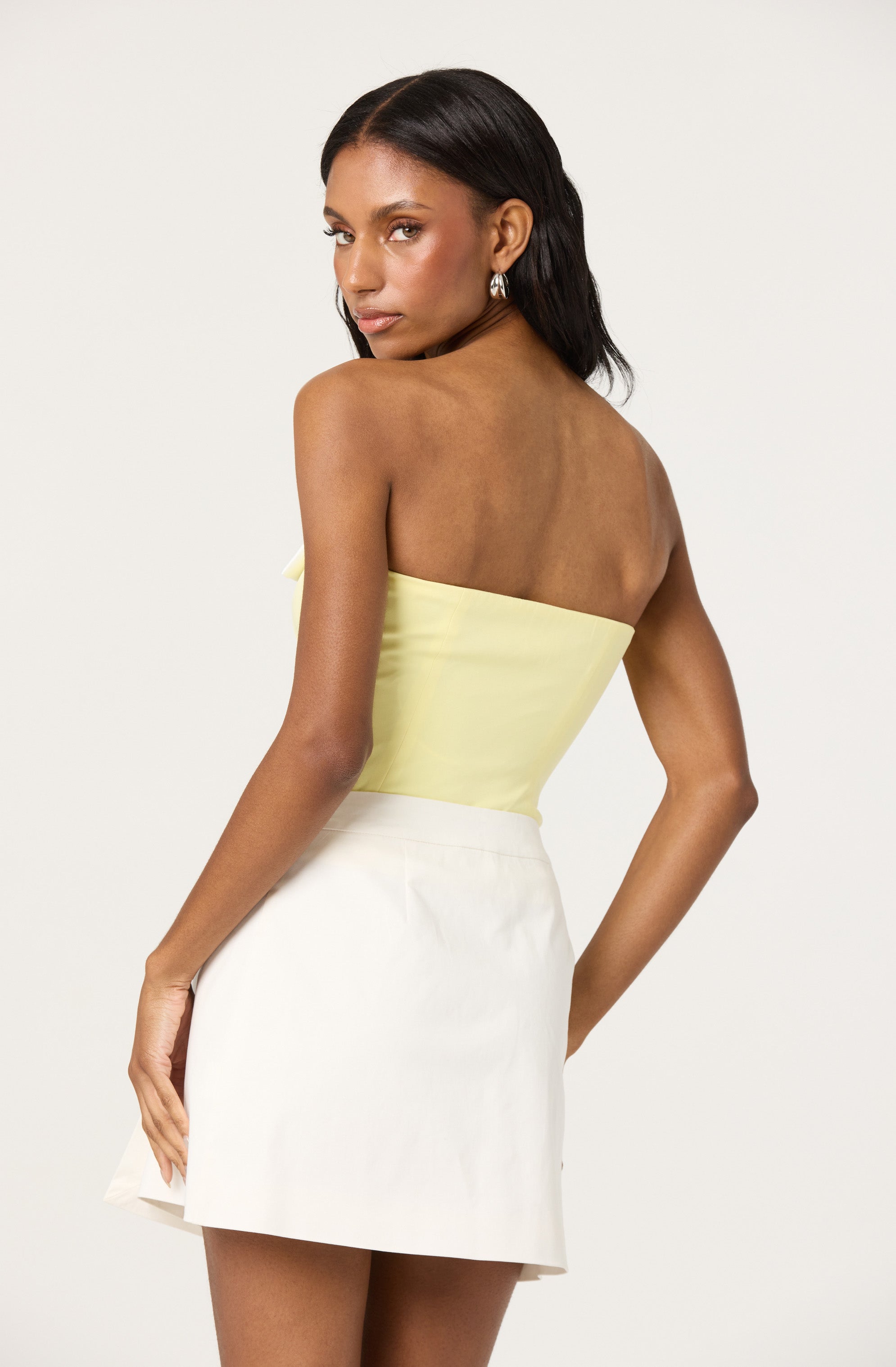 A woman is wearing a strapless, pale yellow satin top and a high-waisted white skirt. She has long, dark hair and is accessorized with silver hoop earrings. She is posing with her back partially turned, looking over her shoulder.