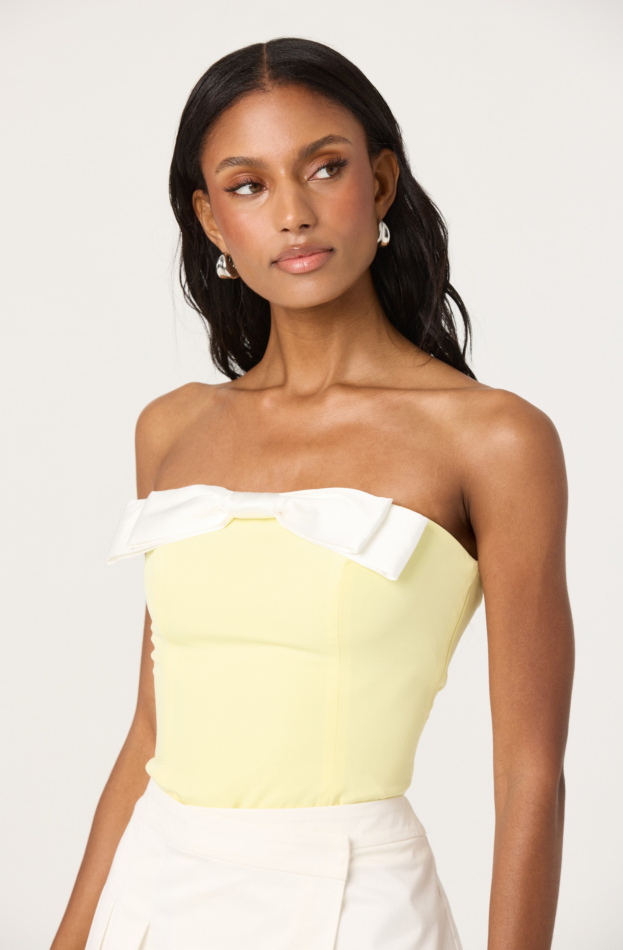 A woman is wearing a strapless, pale yellow top with a large white bow detail across the chest. She pairs it with white high-waisted bottoms. She has dark, wavy hair and is accessorized with large, silver hoop earrings.