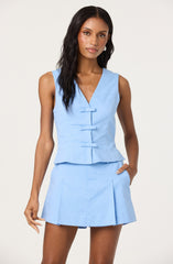 A woman is wearing a sleeveless, light blue two-piece outfit. The top features a V-neckline with three fabric-covered buttons down the front and a peplum hem. The matching high-waisted skirt has pleats and falls above the knee.