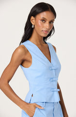 A woman is wearing a sleeveless light blue top with a V-neckline. The top features three fabric-covered toggle closures down the front and has small side slits at the hem. She is also wearing matching light blue pants with her hand resting in her pocket.