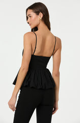 A woman is wearing a black sleeveless top with thin spaghetti straps and a ruffled peplum detail at the waist. The top is fitted around the torso, and the ruffles create a voluminous effect at the hip. She pairs it with matching black pants. She has long, dark hair and is accessorizing with gold hoop earrings.