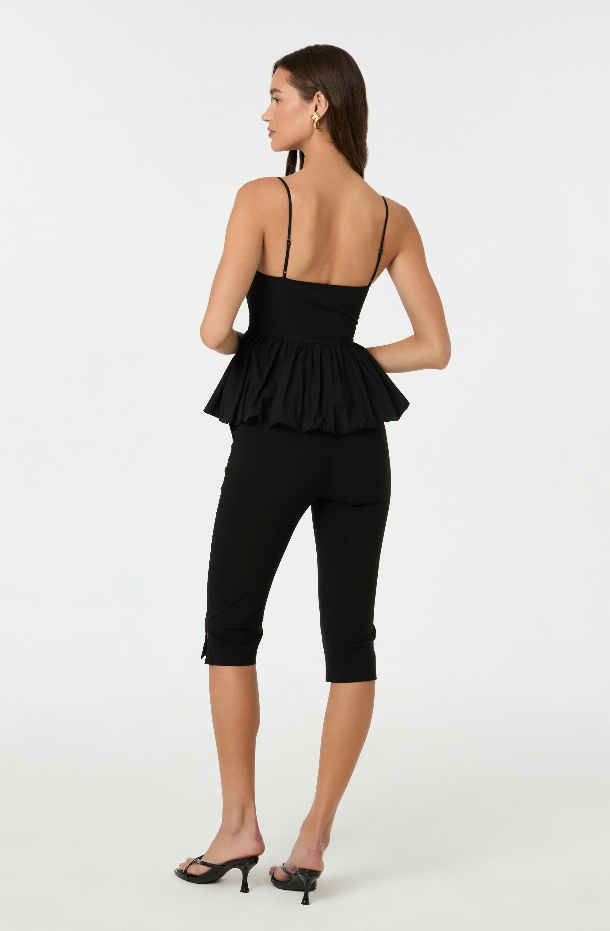 A woman is wearing a black sleeveless top with thin spaghetti straps and a fitted bodice that features a ruffled peplum hem. She pairs it with black cropped pants that fit snugly and end just below the knees. She completes the outfit with black open-toe high-heeled sandals.