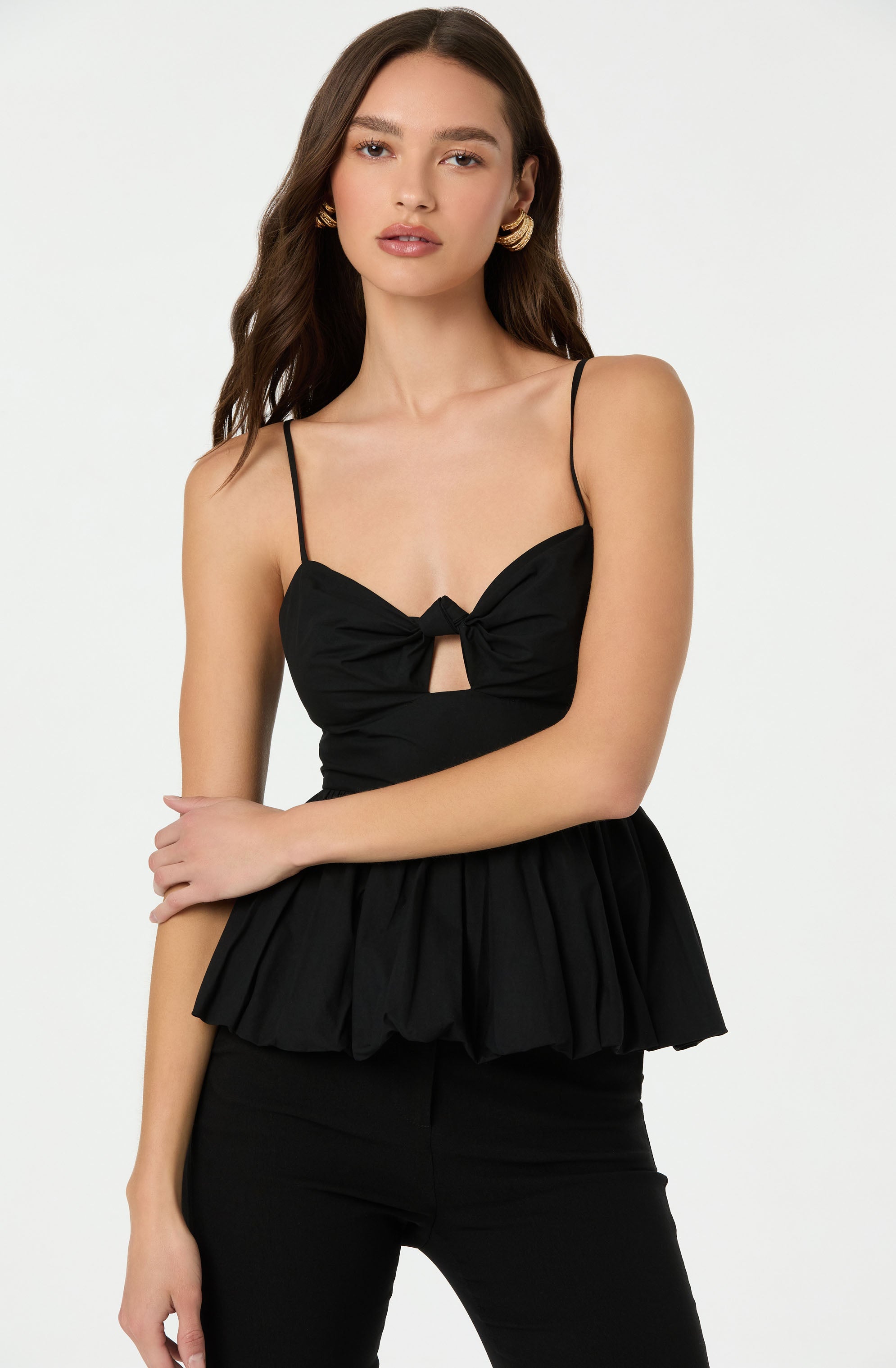 A young woman is wearing a black sleeveless top with thin spaghetti straps. The top features a bow detail at the center with a small cutout underneath. The fabric of the top is gathered below the bow, creating a peplum style. She pairs it with black pants. The woman has long, wavy brown hair and is accessorized with gold hoop earrings.