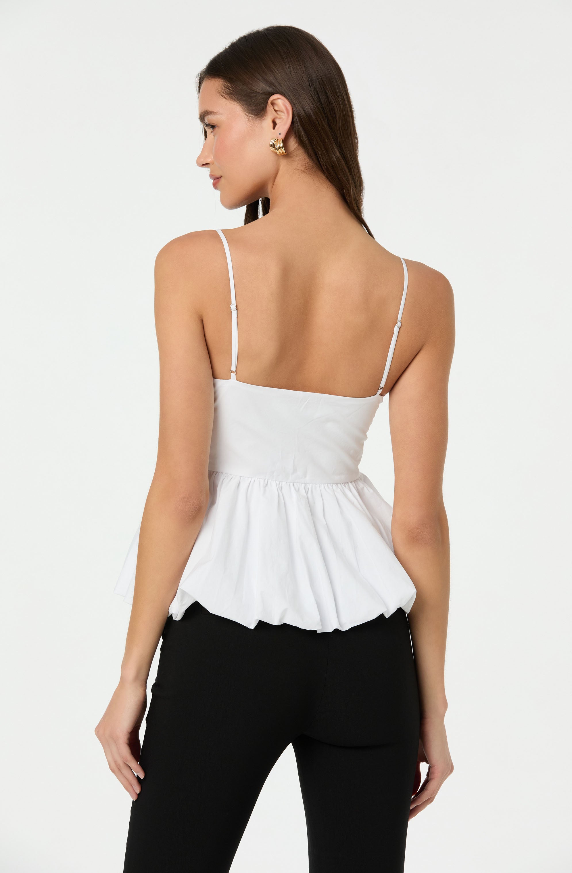 A woman is wearing a white sleeveless top with thin spaghetti straps and a fitted bodice that flares out into a ruffled, peplum-style hem. She is also wearing black pants.