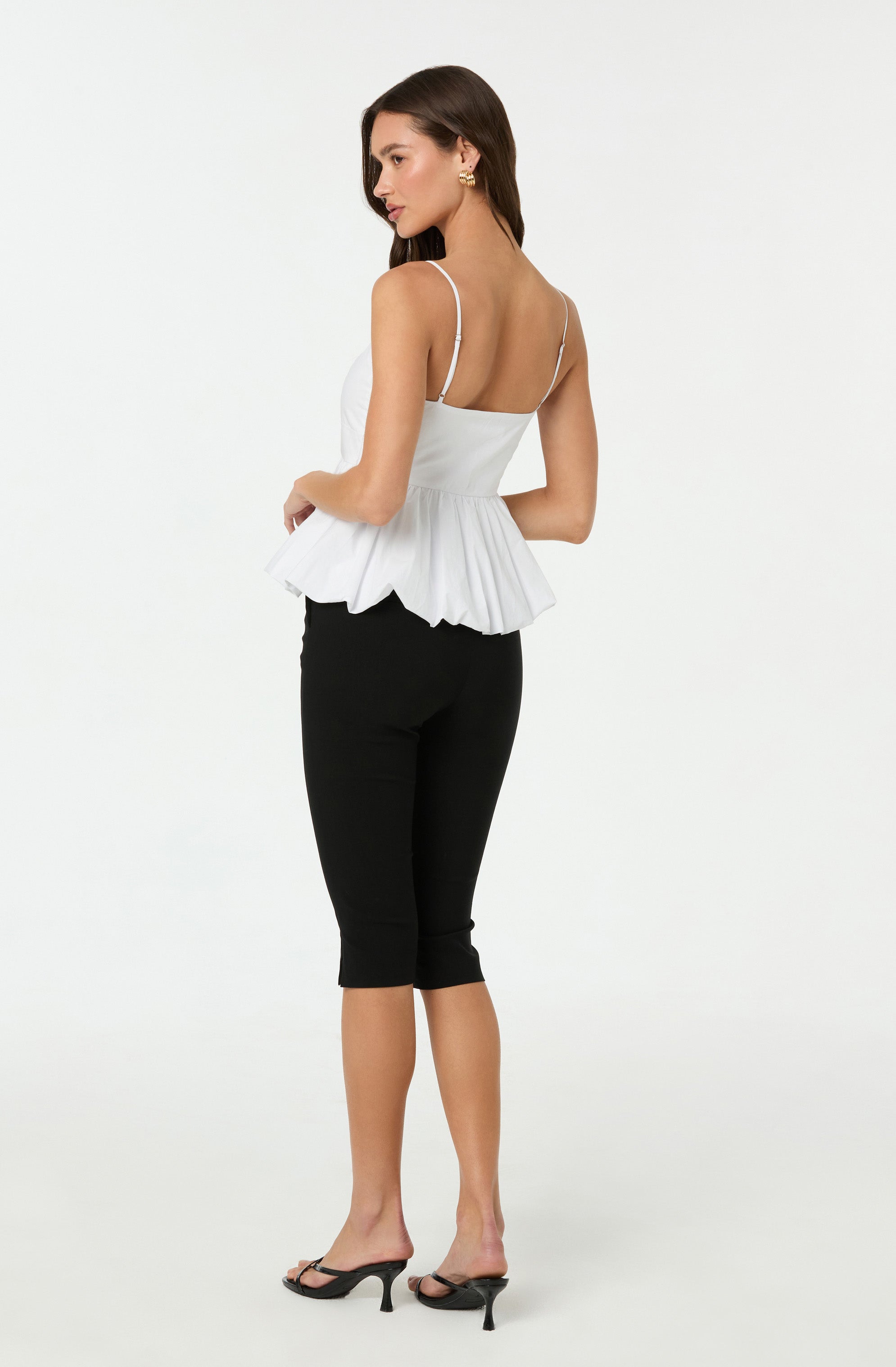 A woman is wearing a white sleeveless top with thin spaghetti straps and a ruffled hem, paired with black cropped pants. She has on black high-heeled sandals with ankle straps.