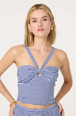 A woman is wearing a blue and white checkered sleeveless top with straps that crisscross over her shoulders, forming a bow-like detail at the front. The top has a fitted design and a small cutout at the center of the chest. She is also wearing matching checkered shorts.
