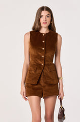 A woman is wearing a sleeveless, brown suede vest with a round neckline and gold buttons down the front. The vest has two flap pockets on the front at hip level. She is also wearing a matching brown suede mini skirt. She accessorizes with gold earrings, a gold bracelet, and holds a small brown handbag with a gold chain strap.