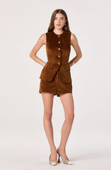 A woman is wearing a sleeveless, brown suede vest with large gold buttons down the front and front pockets. She pairs it with a matching brown suede mini skirt that has a front slit. She is also wearing beige high-heeled shoes with a checkered pattern.