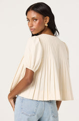 A woman is wearing a cream-colored top with vertical pleats and puffed short sleeves that are gathered at the hem, paired with light blue jeans. She has gold earrings and is standing with her back partially turned, looking over her shoulder.