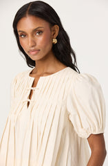 A woman with long, dark hair is wearing an off-white, pleated dress with a round neckline and short, puffed sleeves gathered at the ends. The dress features vertical pleats and a small keyhole opening at the chest, held together by a thin tie. She is also wearing a large, gold hoop earring.