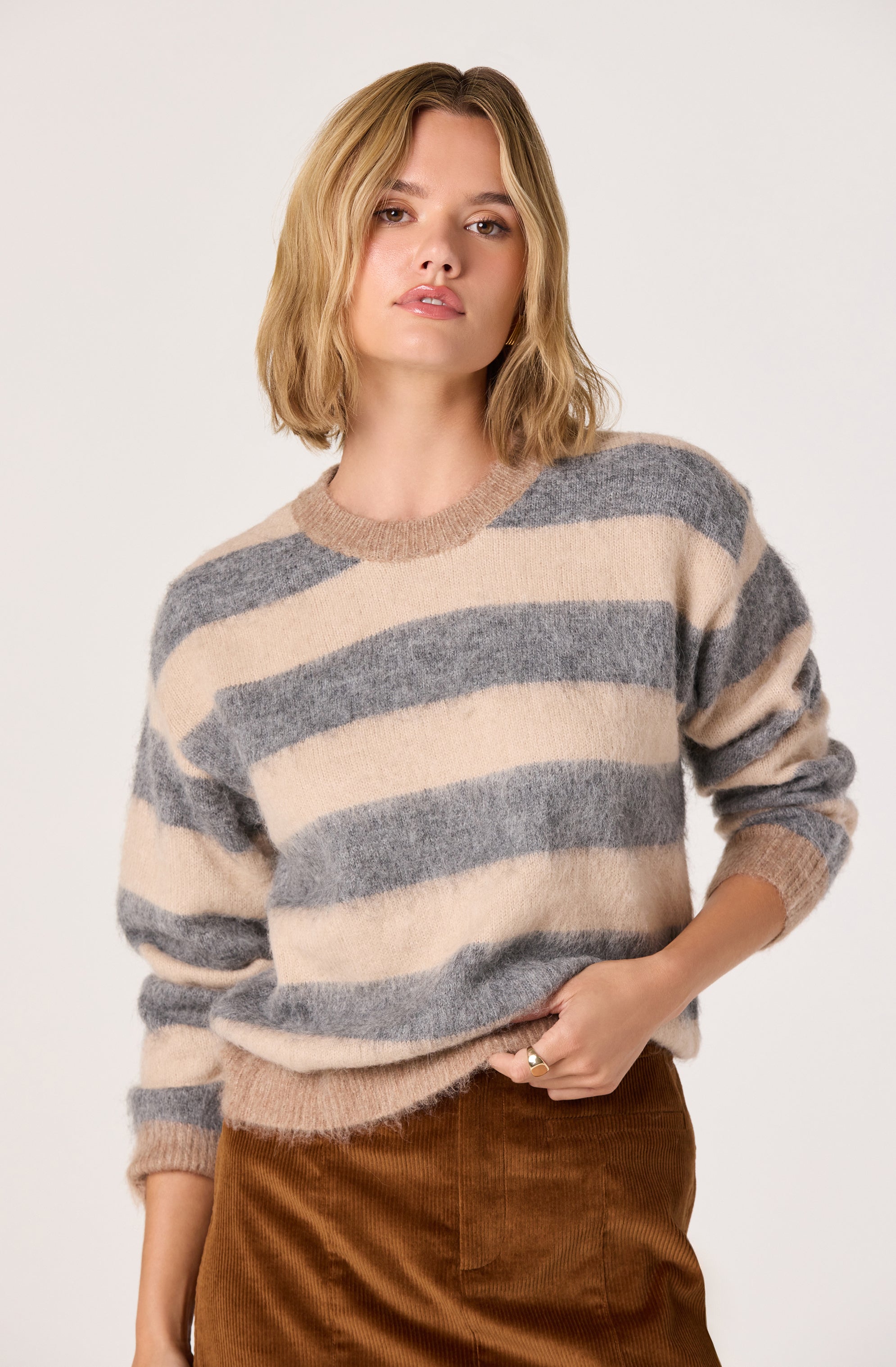 A woman is wearing a cozy, oversized sweater with wide horizontal stripes in neutral tones of beige and gray. The sweater has a round neckline and long sleeves. She is also wearing high-waisted, brown corduroy pants.