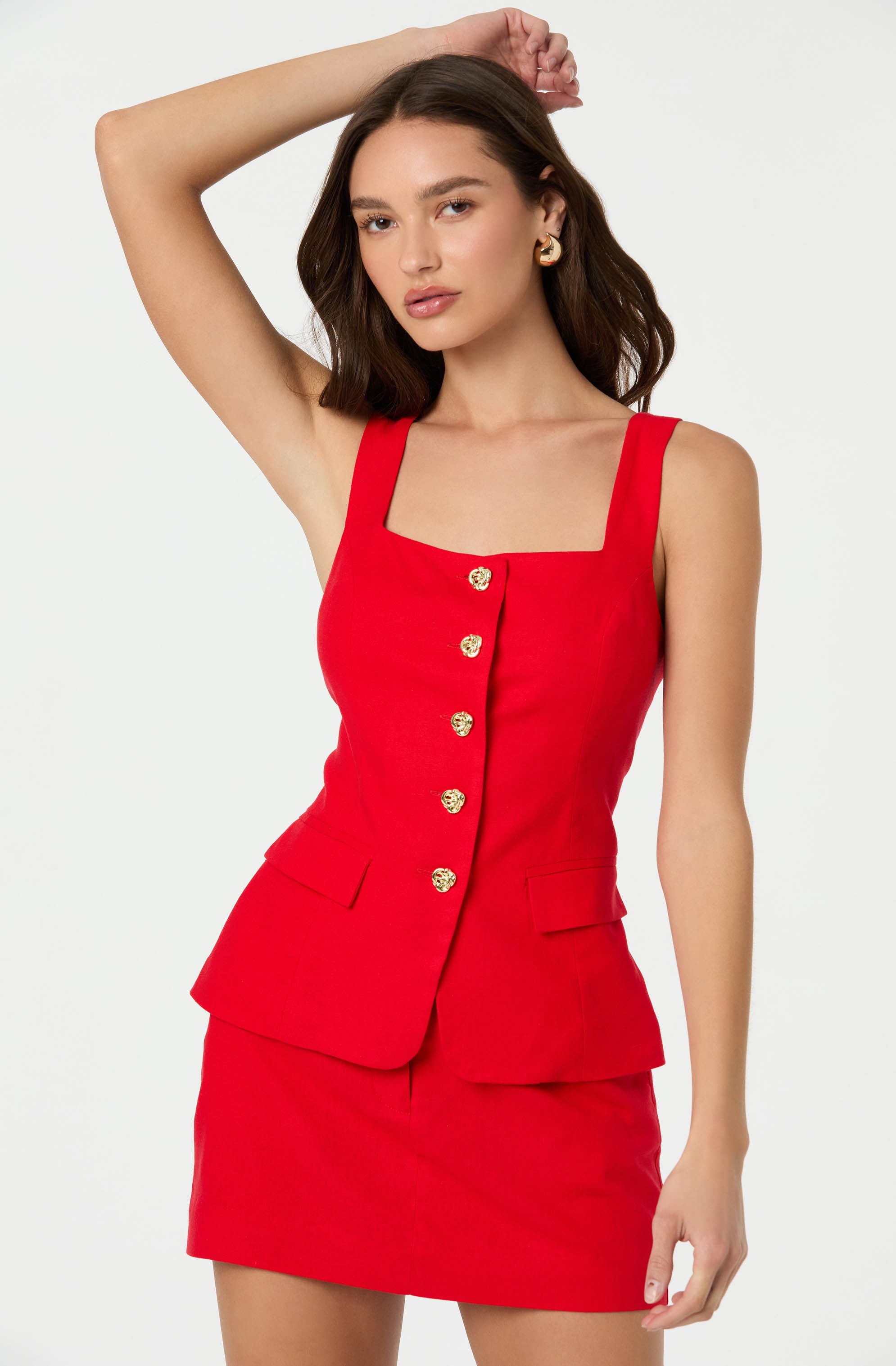 A woman is wearing a sleeveless, fitted red dress with wide shoulder straps. The dress features a square neckline and a front row of decorative gold buttons. The skirt portion of the dress is short and includes a layered, peplum-style overlay with two small flap pockets on the front.